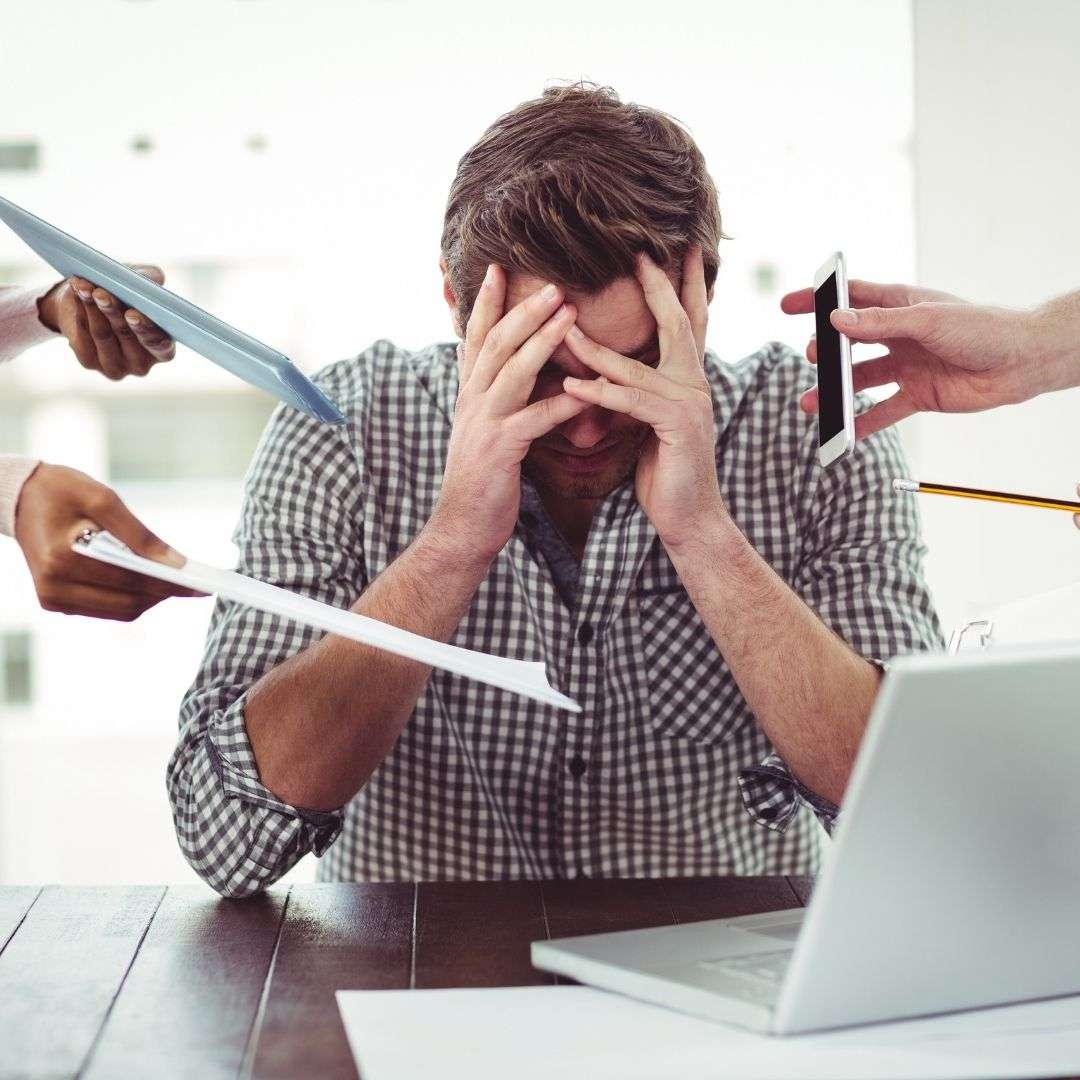 Stressed man at desk overwhelmed by multiple hands presenting documents and a smartphone, illustrating change management challenges.